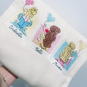 May include: A white tote bag with embroidered images of Cinderella, Belle, and Aurora from Disney. Each image is framed in a Polaroid-style frame with the character's name underneath.