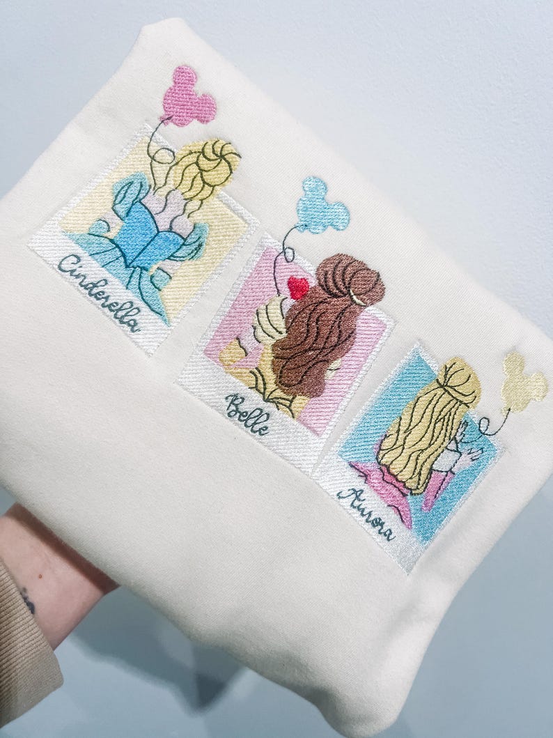 May include: A white tote bag with embroidered images of Cinderella, Belle, and Aurora from Disney. Each image is framed in a Polaroid-style frame with the character's name underneath.