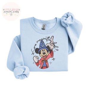 Op de afbeelding: Lichtblauw sweatshirt met een afbeelding van Mickey Mouse in een tovenaarsoutfit met de tekst "Come Imagination". Het ontwerp bevat sterren en een maan. De sweatshirt heeft lange mouwen en een ronde hals.