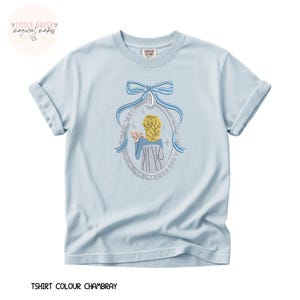 May include: Light blue t-shirt with a detailed embroidered design. The design features a framed image of a person with blonde hair, wearing a blue dress, and a blue bow. The text "TSHIRT COLOUR CHAMBRAY" is at the bottom.