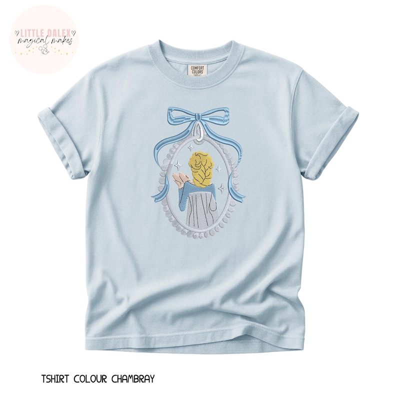 May include: Light blue t-shirt with a detailed embroidered design. The design features a framed image of a person with blonde hair, wearing a blue dress, and a blue bow. The text "TSHIRT COLOUR CHAMBRAY" is at the bottom.