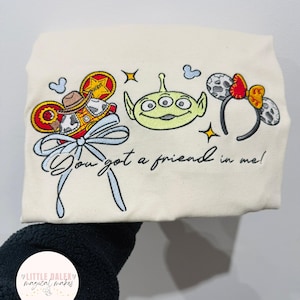 May include: Cream tote bag with embroidered Toy Story characters: Woody, an alien, and Mickey Mouse ears. The phrase "You got a friend in me!" is written in script. The design includes a blue bow and stars.