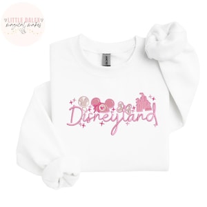 Peut inclure: Sweat-shirt blanc avec un motif rose Disneyland. Le motif comprend le mot "Disneyland" en &eacute;criture cursive, des oreilles de Mickey Mouse, un n&oelig;ud, une sucette et un ch&acirc;teau. Le sweat-shirt porte une &eacute;tiquette "Gildan".