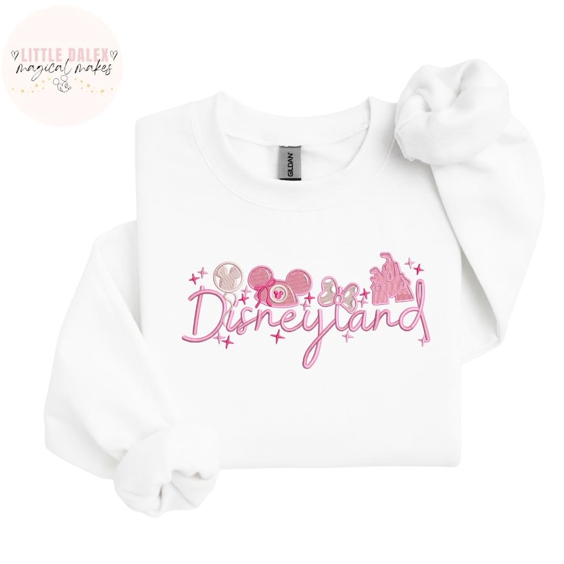Peut inclure: Sweat-shirt blanc avec un motif rose Disneyland. Le motif comprend le mot "Disneyland" en &eacute;criture cursive, des oreilles de Mickey Mouse, un n&oelig;ud, une sucette et un ch&acirc;teau. Le sweat-shirt porte une &eacute;tiquette "Gildan".