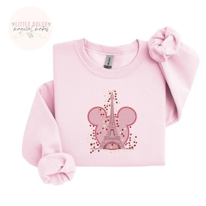 May include: A pale pink sweatshirt with an embroidered design of the Eiffel Tower inside a Mickey Mouse head shape. The design is decorated with red and gold stars. The text "LITTLE DALEX magical makes" is in a circle in the upper left corner.