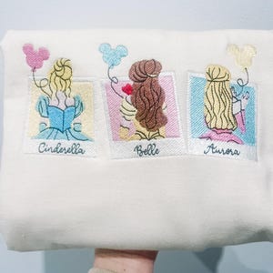 May include: White sweatshirt with embroidered images of Cinderella, Belle, and Aurora from Disney. Each princess is holding a Mickey Mouse balloon and has their name written below their image.