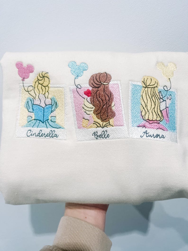 May include: White sweatshirt with embroidered images of Cinderella, Belle, and Aurora from Disney. Each princess is holding a Mickey Mouse balloon and has their name written below their image.