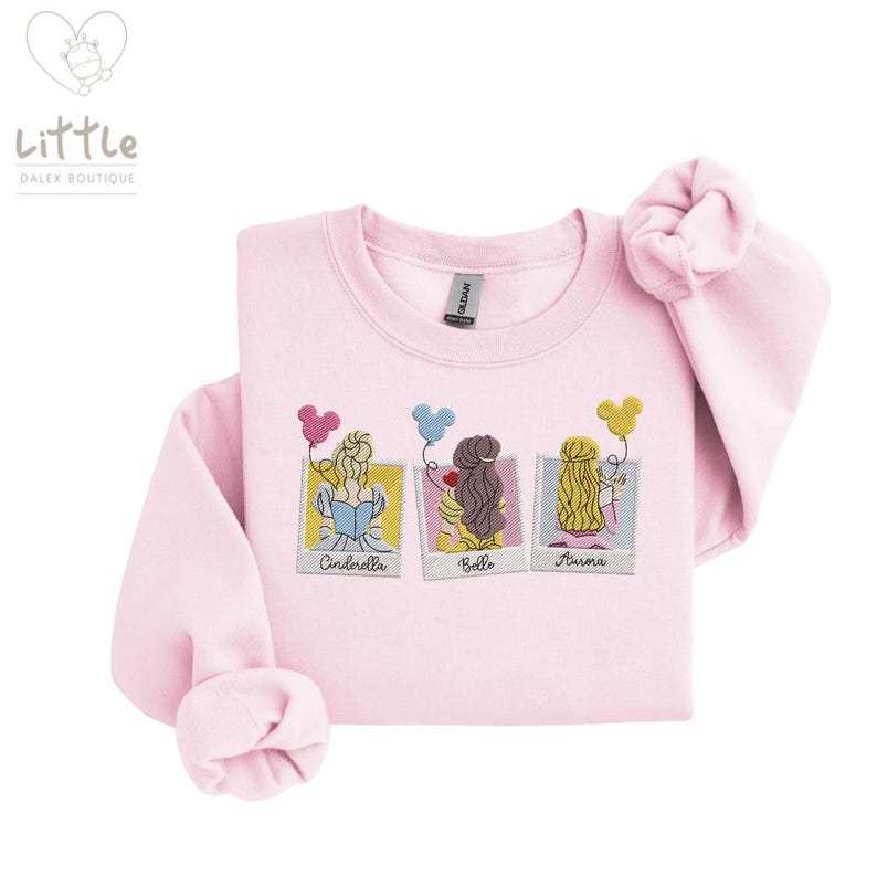 May include: Pink sweatshirt with a graphic of three Disney princesses, Cinderella, Belle, and Aurora, in a photo booth with balloons. The text "Cinderella", "Belle", and "Aurora" is below each princess.