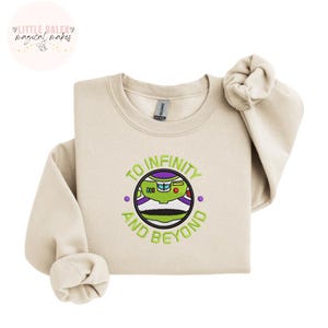 Puede incluir: Sudadera color crema con cuello redondo y mangas largas. Presenta un dise&ntilde;o bordado de Buzz Lightyear con el texto "TO INFINITY AND BEYOND" en verde. La marca "Little Daley Magical Makes" est&aacute; en la esquina superior izquierda.
