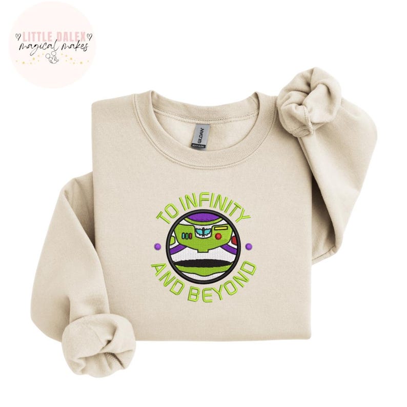 Puede incluir: Sudadera color crema con cuello redondo y mangas largas. Presenta un dise&ntilde;o bordado de Buzz Lightyear con el texto "TO INFINITY AND BEYOND" en verde. La marca "Little Daley Magical Makes" est&aacute; en la esquina superior izquierda.