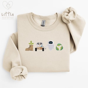 May include: A beige sweatshirt with an embroidered design featuring a robot, a plant, a boot, and a recycling symbol. The design is in shades of brown, green, and white.