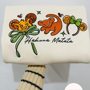 Op de afbeelding: Crèmekleurige sweatshirt met geborduurde Lion King-personages en de zin "Hakuna Matata". Het ontwerp bevat Simba, een leeuwenwelp en muisoren-haarbanden met strikken. De sweatshirt wordt vastgehouden door een persoon die een beige trui met zwarte strepen draagt.