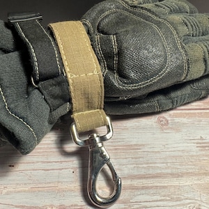 May include: A pair of work gloves in shades of gray and green, with a tan strap and a silver metal clip. The gloves are made of a durable material and appear to be designed for heavy-duty use. The clip is attached to the strap and is designed to secure the gloves.