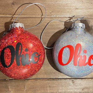 May include: Two Christmas ornaments with glitter. One is red with black lettering that says "Ohio". The other is silver with red lettering that says "Ohio".