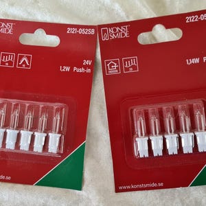 Könnte beinhalten: Zwei rote Verpackungen mit Weihnachtsglühbirnen. Jede Packung enthält fünf kleine, klare Glühbirnen mit weißen Sockeln. Die Verpackungen tragen die Aufschrift "KONST SMIDE" und "INDOOR". Eine Packung ist 24V, 1,2W Push-in, die andere 12V, 1,14W Push-in.