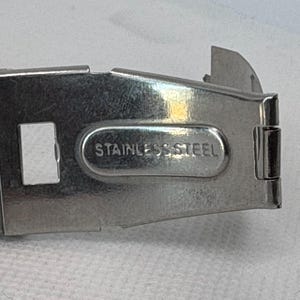 Pode incluir: Uma pulseira de rel&oacute;gio de metal prateado com uma fivela de a&ccedil;o inoxid&aacute;vel. A fivela tem uma abertura retangular e as palavras "Stainless Steel" gravadas nela.