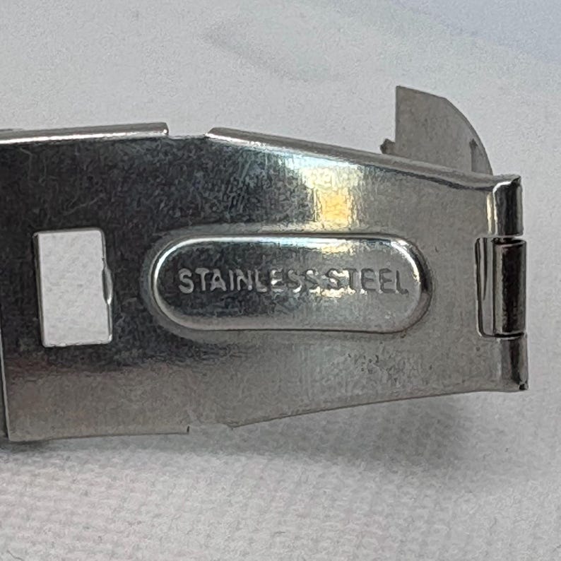 Pode incluir: Uma pulseira de rel&oacute;gio de metal prateado com uma fivela de a&ccedil;o inoxid&aacute;vel. A fivela tem uma abertura retangular e as palavras "Stainless Steel" gravadas nela.