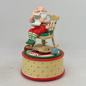 Può includere: Una statuina musicale a tema natalizio di Babbo Natale seduto su una sedia a dondolo, che legge una lista. La statuina è su una base rotonda con un bastoncino di zucchero e una palla blu e gialla. La base ha un motivo giallo con accenti rossi.