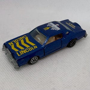 May include: A blue die-cast Lincoln Continental Mark V toy car. The car features yellow and black detailing, including the word "LINCOLN" and an eagle emblem. The car has black wheels and a tan interior.