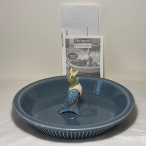 Puede incluir: Una fuente para pastel de cerámica azul con una figura decorativa de pájaro en el centro. El pájaro es verde, azul y blanco. Un folleto de recetas de Pfaltzgraff Perfect Pie Plate está detrás del plato.