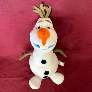 Può includere: Un peluche a forma di pupazzo di neve con corpo bianco, dettagli di bottoni neri e naso a carota. Il pupazzo di neve ha un viso sorridente con occhi blu, bocca blu e braccia e capelli di ramoscelli. Il giocattolo è su uno sfondo rosso.