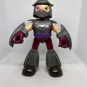 May include: A plastic action figure of Shredder from Teenage Mutant Ninja Turtles. The figure has a gray helmet, armor, and boots, with a purple belt and accents. The figure's face is visible, with a pink and yellow mask.