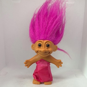 May include: A vintage troll doll with a tan face, large black eyes, and a cheerful smile. The doll has bright pink, upright hair and wears a pink fabric garment. The arms are outstretched, and the feet are visible.