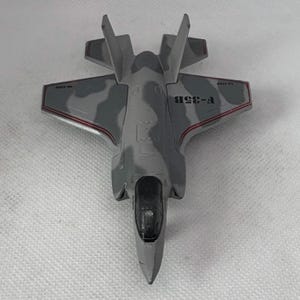 May include: A gray toy airplane with a camouflage pattern on the wings and body. The model plane has the text "F-35B" printed on the side. The plane has a black cockpit and red trim.