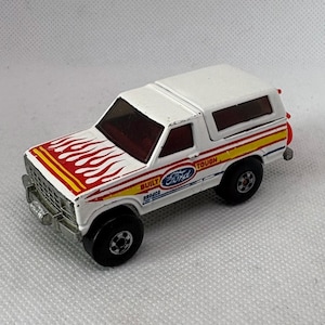 May include: A white Ford Bronco toy truck with red, yellow, and blue accents. The hood features a flame design. The words "Ford", "Built Bronco", and "Tough" are visible on the side.