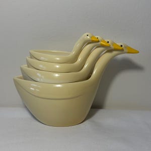 May include: A set of four nested measuring cups shaped like geese, featuring a smooth cream-coloured finish with bright yellow beaks and small black eyes. The cups are stacked together, with each goose head extending outward to form a handle.