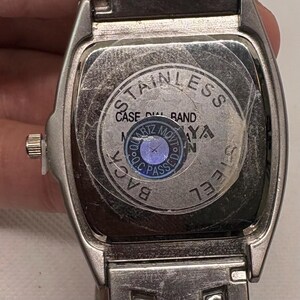 Pode incluir: Um rel&oacute;gio de pulso de a&ccedil;o inoxid&aacute;vel prateado com um mostrador retangular. O mostrador do rel&oacute;gio tem um fundo preto com texto branco que diz "STAINLESS CASE DIAL BAND BACK STEEL SIDE". O mostrador do rel&oacute;gio tamb&eacute;m tem um adesivo redondo azul com texto branco que diz "QUARTZ MOVT O.C. PASSED".