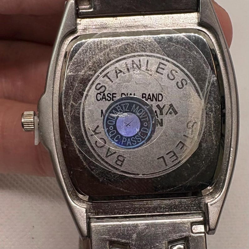 Pode incluir: Um rel&oacute;gio de pulso de a&ccedil;o inoxid&aacute;vel prateado com um mostrador retangular. O mostrador do rel&oacute;gio tem um fundo preto com texto branco que diz "STAINLESS CASE DIAL BAND BACK STEEL SIDE". O mostrador do rel&oacute;gio tamb&eacute;m tem um adesivo redondo azul com texto branco que diz "QUARTZ MOVT O.C. PASSED".