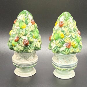 May include: A pair of ceramic salt and pepper shakers. Each shaker is shaped like a Christmas tree with green leaves, yellow fruit, and brown pinecones. The base is a light green urn shape with a decorative design.