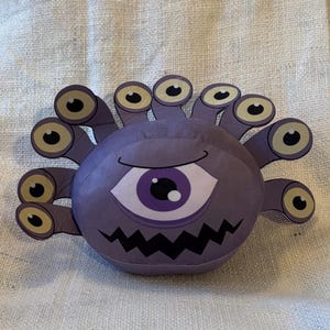 May include: Purple plush toy with multiple eyes and a large central eye. The toy has a jagged mouth and is shaped like a sphere.