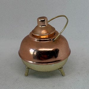 May include: A small, copper-colored metal teapot with a gold-colored base and handle. The teapot has a rounded body, a lid, and three small legs. The handle is a simple, curved wire. The teapot is likely a decorative item or a miniature collectible.