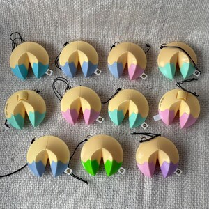 May include: A selection of fortune cookie keyrings. Each keyring has a yellow outer shell and a colourful interior, with options including blue, pink, green, and purple. They have a black string and a small tag attached.