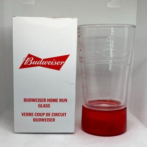 May include: A white box with the Budweiser logo in red and the text "Budweiser Home Run Glass" and "Verre Coup de Circuit Budweiser". A clear glass with a red base and the Budweiser logo etched on the side.