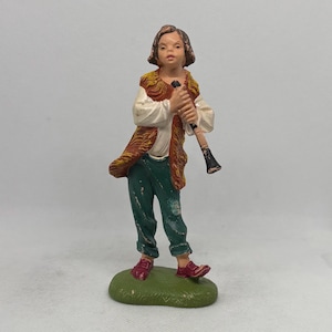 Puede incluir: Figura de una persona tocando un instrumento de viento. La figura viste camisa blanca, pantalón verde y chaleco marrón. La figura se encuentra sobre una base verde y sostiene un instrumento negro y marrón. La figura tiene cabello castaño y zapatos rojos.