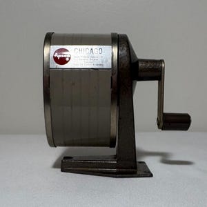 May include: A vintage manual pencil sharpener with a dark metallic base and a cylindrical body. The side features a label reading Apsco Chicago, Apsco Products (Canada) Ltd., Toronto, Ontario, Canada, Type 2A Cutter Assembly, and a hand crank.