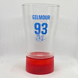May include: A clear glass with a red base. The glass features the word "GILMOUR" in blue, the number "93", and a signature. The glass is cylindrical and is likely a drinking glass.