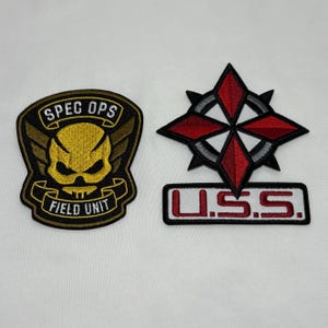 May include: Two embroidered patches. One is a black and gold Spec Ops Field Unit patch with a skull design. The other is a red and black star-shaped patch with the letters U.S.S. in a white rectangle.