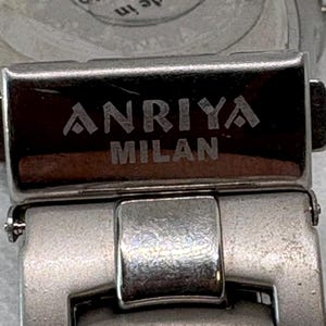 Pode incluir: Close-up de uma pulseira de rel&oacute;gio prateada com as palavras "ANRIYA MILAN" gravadas na fivela.