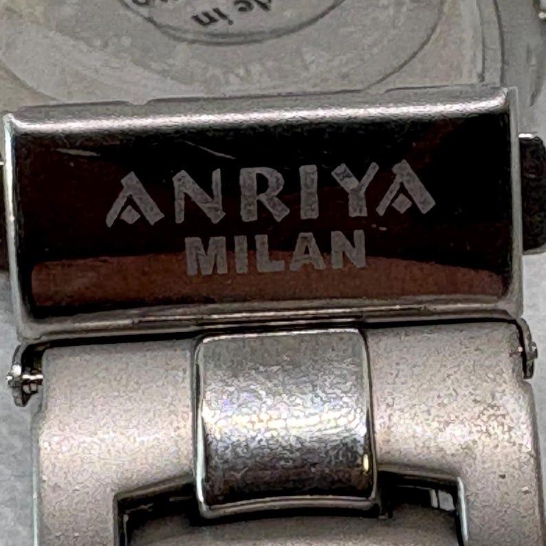 Pode incluir: Close-up de uma pulseira de rel&oacute;gio prateada com as palavras "ANRIYA MILAN" gravadas na fivela.