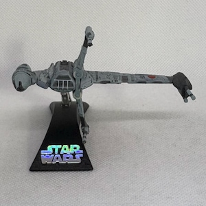 May include: A gray Star Wars B-Wing fighter model on a black stand. The model has detailed wings and a cockpit. The base features the Star Wars logo in iridescent colors. The fighter is a collectible item.