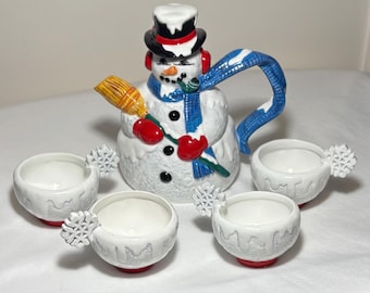 May include: A whimsical snowman-shaped teapot with a black top hat, red earmuffs, and a blue scarf. The set includes four white teacups with snowflake handles and red bases. The teapot holds a broom.