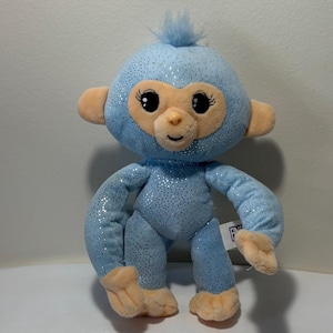 May include: A plush blue monkey with glitter accents. The stuffed animal has a light brown face, ears, hands, and feet. The monkey has large black eyes with eyelashes and a small tuft of blue hair on top of its head.
