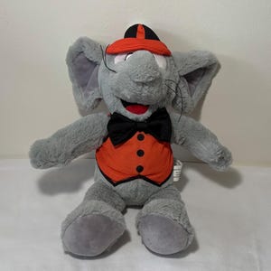 Puede incluir: Un peluche gris con forma de elefante que lleva un chaleco naranja con botones negros, una pajarita negra y un sombrero rojo y negro. El elefante tiene orejas grandes, boca roja y bigotes negros.