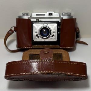May include: A vintage Fex Elite camera with a brown leather case. The camera is silver and black with a lens and dials. The case is open, revealing the camera. The camera strap is attached to the side of the case.