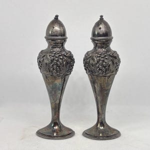 Può includere: Un paio di saliere e pepiere antiche in argento. Ogni shaker presenta un design dettagliato a grappolo d'uva attorno al corpo e una parte superiore conica perforata. Gli shaker hanno una forma sottile, a forma di vaso e una finitura ossidata.