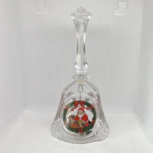 May include: A clear glass Christmas bell with a long handle and a decorative image of Father Christmas inside a wreath. The bell has a diamond-cut pattern around the base and a clear, pointed handle.